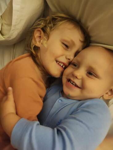 A imagem mostra duas crianças pequenas, um menino e uma menina, deitados em uma cama, abraçados, com uma marca d'água no centro.