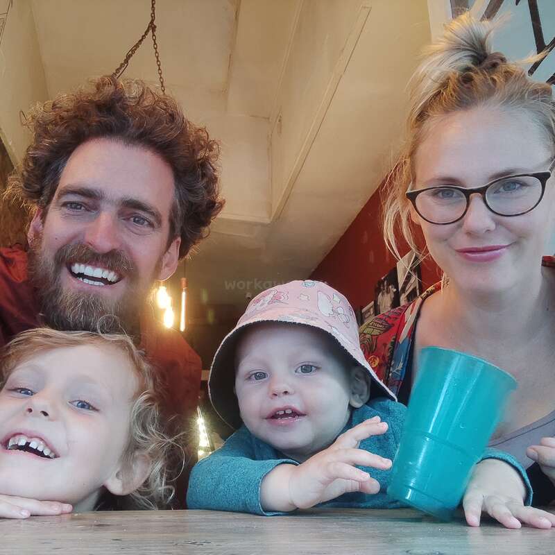 A imagem mostra uma família de quatro pessoas posando para uma foto, com duas crianças e dois adultos, em uma sala com paredes brancas e uma parede vermelha ao fundo.