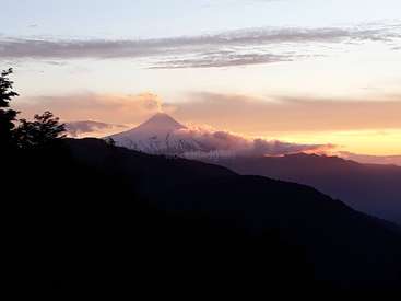The image depicts a majestic mountain with snow at its peak, partially shrouded in clouds, set against a vibrant sky with hues of orange and yellow at sunset.