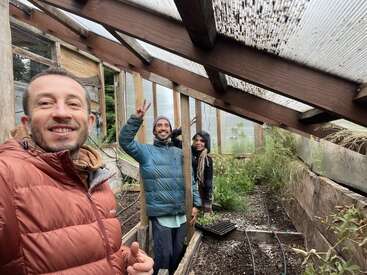Three people smiling in a greenhouse, dressed warmly. They are surrounded by plants, wooden beams, and clear roof panels. The atmosphere is cheerful and relaxed.