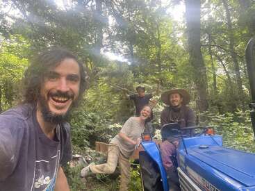 Quatro pessoas felizes aproveitam um dia ensolarado na floresta, posando com um trator azul. Parecem estar trabalhando juntas, sorrindo, cercadas por uma vegetação exuberante.