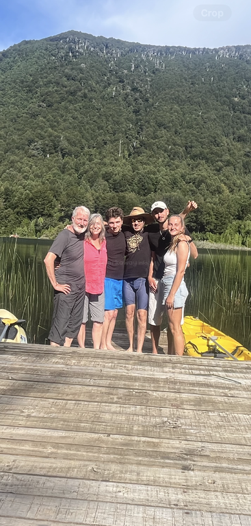 Six people stand close together on a wooden dock, smiling, with a yellow kayak, green lake, and forested mountain under a blue sky in the background.