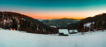 A serene winter landscape featuring snow-covered hills, scattered wooden cabins, and dense evergreen forests at sunset, with a beautiful gradient sky fading into warm colors.