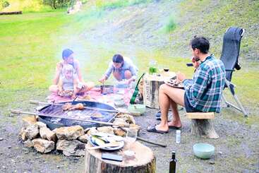 A family enjoys a picnic outdoors, sitting on blankets and makeshift seats around a campfire, grilling food, eating, and relaxing in a grassy, scenic area.