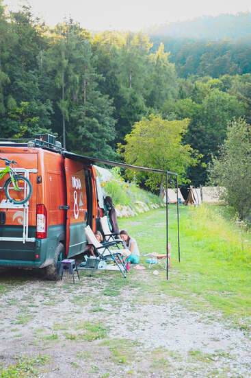 A bright orange camper van parked amid lush greenery, with two people relaxing on chairs under an awning, enjoying a peaceful outdoor camping experience.
