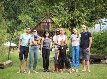 A group of adults and children are standing outside on grass, smiling together. Behind them are trees, a rustic wooden building, and lush greenery.