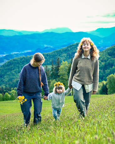 Three people—two adults and a child—hold hands while walking through a grassy field. They wear casual clothes and flower crowns, surrounded by beautiful mountains and greenery.