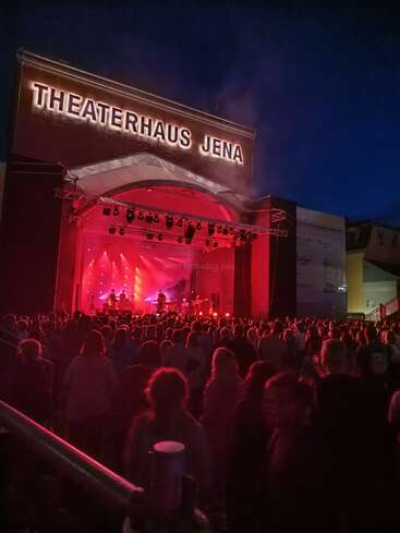 Abends versammelt sich eine große Menschenmenge vor dem Theaterhaus Jena und genießt eine Live-Musikdarbietung auf einer hell erleuchteten Bühne mit leuchtend roter Beleuchtung.