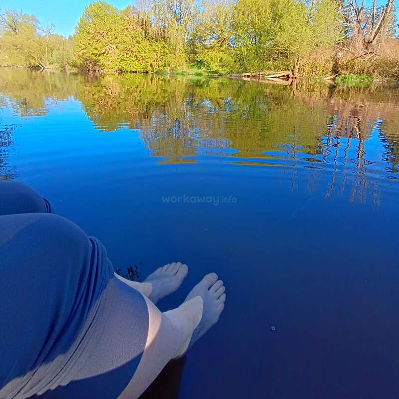 Zwei Menschen sitzen an einem ruhigen blauen See, tauchen ihre Füße ins Wasser und sind umgeben von üppig grünen Bäumen, die sich wunderschön auf der ruhigen Oberfläche spiegeln.