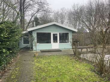 A small, light-blue wooden cabin with large windows sits amidst bare trees. There is a patchy lawn, potted plant, and wooden fencing in the foreground.