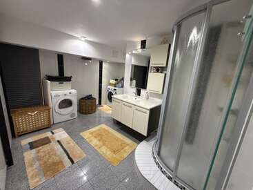 Salle de bains spacieuse équipée d'une douche moderne, d'un meuble-lavabo à double vasque, d'un lave-linge, d'un sèche-linge, d'un panier à linge en osier, d'une armoire de rangement et de trois tapis beiges posés sur un sol carrelé brillant.