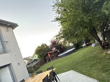 Une scène de jardin montre une maison moderne, une table en bois avec des chaises noires, une grande pelouse verte, des arbres, une piscine extérieure, un abri de jardin, le tout dans une atmosphère détendue et paisible.