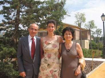 Três pessoas posam ao ar livre num dia ensolarado. Um senhor mais velho de terno, uma mulher com um vestido florido e outra mulher estão juntos, sorrindo.