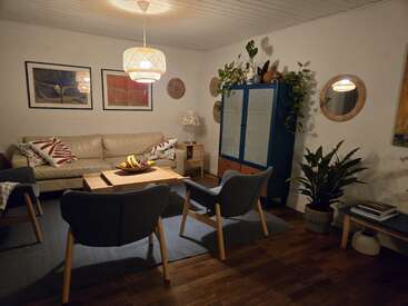 A cozy living room with a beige sofa, two gray chairs, plants, blue cabinet, warm lighting, wall art, mirrors, and a wooden coffee table with fruit.