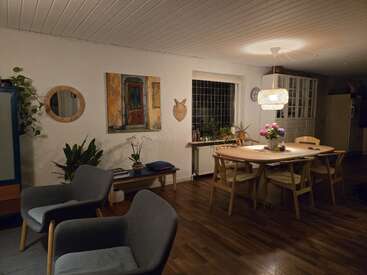 A cozy dining and living area featuring wooden floors, a large table with chairs, modern armchairs, plants, wall art, and a hanging lamp for warm lighting.