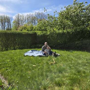 A father and child sit on a blue blanket in a grassy backyard, accompanied by a dog. Trees and shrubs surround them, creating a peaceful, sunny scene.