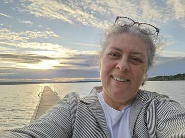 A woman takes a selfie on a wooden pier over a serene lake at sunset, wearing glasses on her head, smiling, with dramatic clouds in the sky.