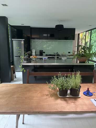 This modern kitchen features black cabinets, a stainless steel refrigerator, and a wooden table with potted plants. Large windows provide natural light and lush views.