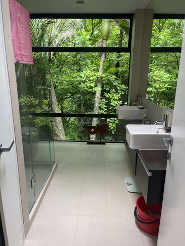 This modern bathroom has large windows overlooking a lush green forest, a glass shower, white sinks, a pink towel, and a red bucket on the floor.