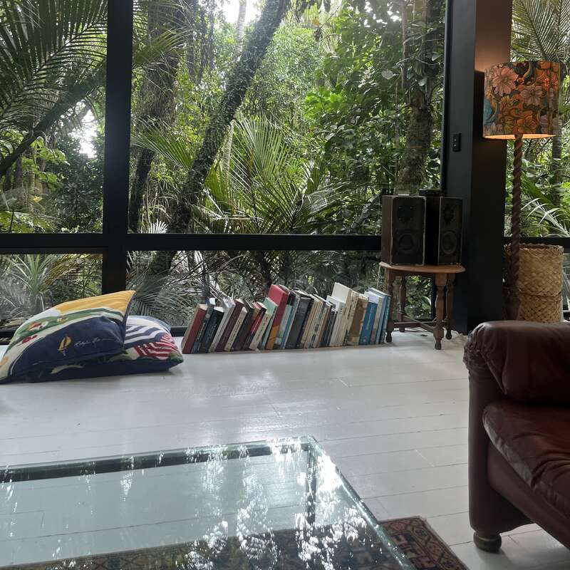 Cozy living room with large glass windows overlooking lush greenery, white floors, books stacked on the floor, colorful pillows, a vintage lamp, and speakers.