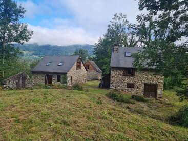 A imagem retrata uma cena rural serena com três casas de pedra com telhados cinza, aninhadas entre árvores e um exuberante campo gramado, em um pitoresco cenário montanhoso.