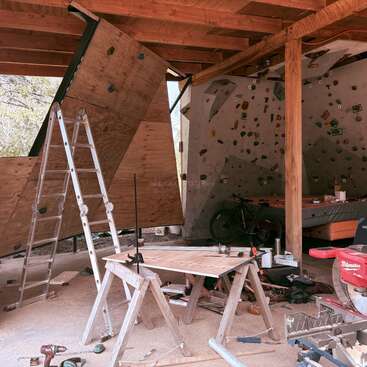 This image depicts an indoor climbing wall under construction. Tools, a workbench, and a ladder are scattered, with a bicycle and mattress visible in the background.