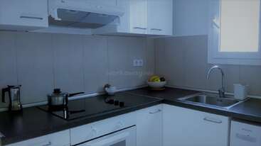 Una cocina moderna y diáfana con armarios blancos, cocina eléctrica, cacerola, cafetera, frutero, fregadero, ventana y una iluminación tenue que crea un ambiente tranquilo y ordenado.