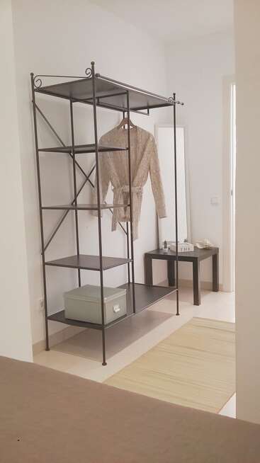 Una habitación minimalista cuenta con un armario metálico abierto en el que hay una bata beige, una caja de almacenamiento verde, un espejo, una mesa negra y una alfombra beige.