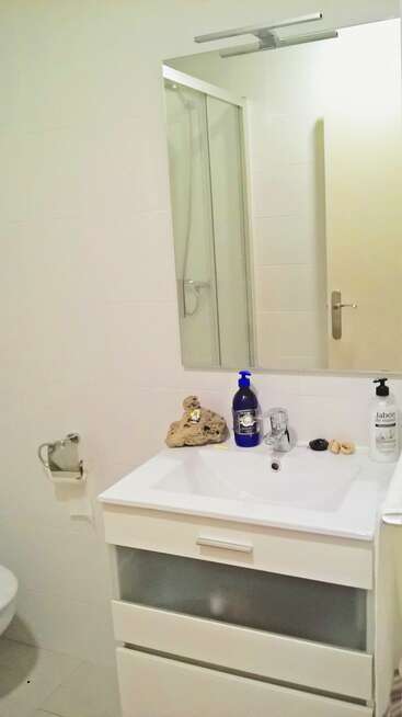Esta imagen muestra un cuarto de baño moderno y minimalista con un mueble de lavabo blanco, un espejo, jabón de manos, una botella azul, una piedra, conchas marinas, una ducha, un inodoro y un toallero.