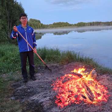 Une personne vêtue d'une veste bleue se tient près d'un lac paisible, un râteau à la main, s'occupant d'un grand feu de camp lumineux, entourée de verdure et d'une eau calme.