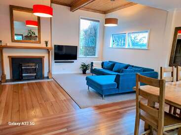 A cozy living room with a blue sectional sofa, wooden floors, fireplace, wall-mounted TV, dining table, large windows, modern lighting, and neutral walls.