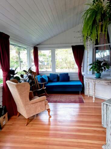 A cozy sunroom with wooden floors, white paneled ceiling, blue sofa, brown armchair, lush plants, large windows, red curtains, and natural light illuminating the space.