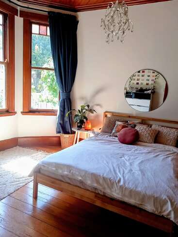 This cozy bedroom features a wooden bed, fluffy pillows, round mirror, blue curtains, chandelier, plant, and large windows allowing natural sunlight to fill the room.