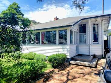 This image shows a charming white house with large windows, a small porch, lush green garden, brick pathway, and trees, under a bright blue sky.