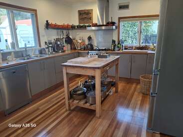 This cozy kitchen features wooden floors, an island with pots, gray cabinets, two windows, stainless appliances, open shelves, and neatly arranged utensils, creating a homely atmosphere.