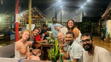 Un groupe d'amis est assis autour d'une table et déguste des boissons et de la nourriture le soir, en souriant et en posant pour l'appareil photo dans un cadre intérieur confortable et décontracté.