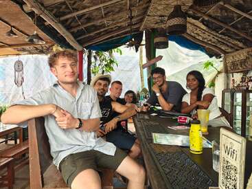 Six amis sont assis dans un bar rustique au toit de bambou, discutant et souriant, des boissons sur la table, créant une atmosphère confortable, détendue et multiculturelle.