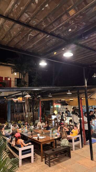 A cozy, lively outdoor restaurant at night features groups of people dining, talking, and relaxing under warm lighting, with tropical plants and a welcoming, casual atmosphere.