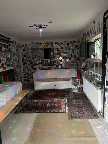 This room features colorful disco lights, decorated walls with photos and CDs, Persian rugs, a white bench bed, a bench, and a cozy, artistic vibe.