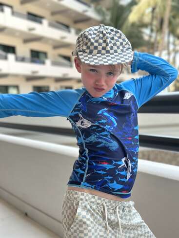 Un niño pequeño posa con confianza, con un sombrero a cuadros, pantalones cortos a juego y una camiseta azul con motivos de tiburones. El fondo muestra un edificio moderno y palmeras.