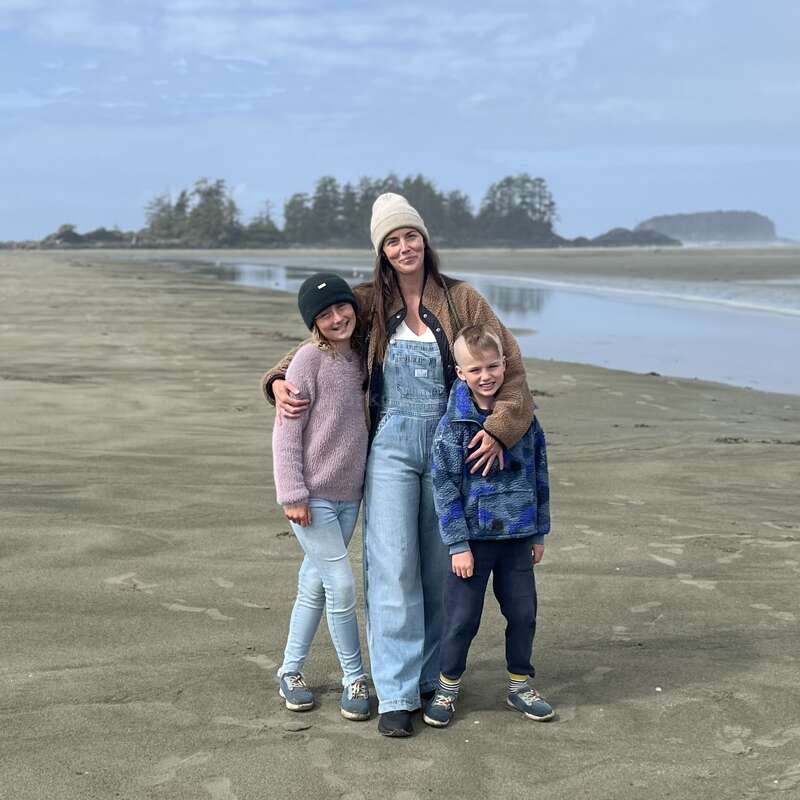Una mujer y dos niños están juntos en una amplia playa de arena, sonriendo. Visten ropa informal y abrigada. Al fondo se ven árboles, agua y un cielo nublado.
