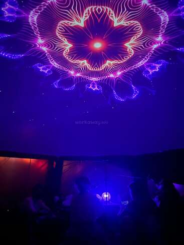 A group of people sit in a dark room under a ceiling with vibrant purple and pink kaleidoscopic light patterns, creating a mesmerizing, immersive, and relaxing atmosphere.