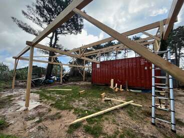 A wooden frame structure is being built outdoors near a red shipping container. Construction materials, a ladder, and scattered wood pieces are visible on muddy ground. Trees surround.