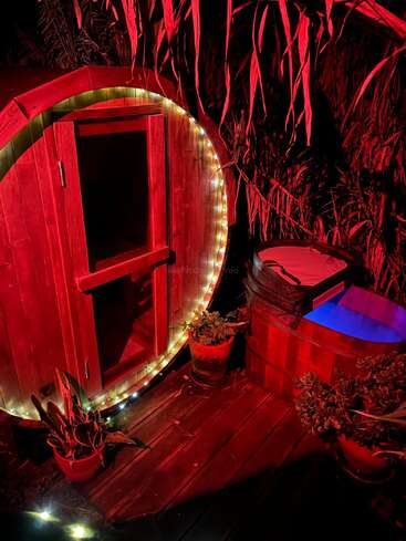 A cozy wooden sauna with string lights stands beside a small hot tub, surrounded by lush plants and bathed in dramatic red lighting at night.