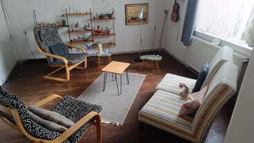 This cozy living room features patterned armchairs, a striped sofa, a small coffee table, shelves with various items, a guitar on the wall, and soft natural light.