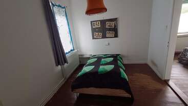 A small, minimally furnished bedroom features a single bed with a geometric green and black blanket, orange hanging lamp, curtained window, wall art, and wooden floor.