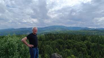 An older man stands on a hilltop, hands on hips, overlooking a lush green forest and distant mountains under a partly cloudy, dramatic sky.
