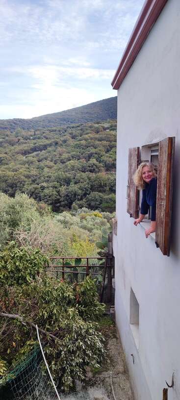 Eine Frau steht vor einem Haus, trägt ein weißes Hemd und eine Jeans, hat die Hände in die Hüften gestemmt und posiert für ein Foto. Sie hat blondes Haar und steht vor einem Haus.
