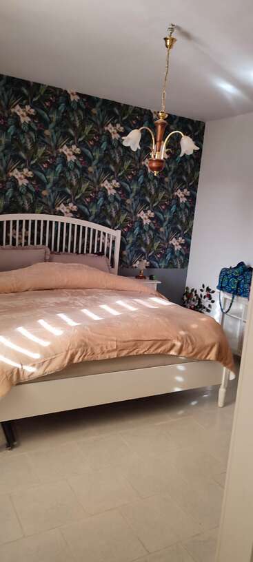 Das Bild zeigt ein Schlafzimmer mit Bett, Nachttisch und Lampe, mit einer weißen Bettdecke und Kissen sowie einer weißen Kommode mit Spiegel vor einer weißen Wand.