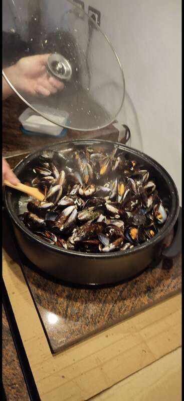 Une grande casserole de moules mijote sur la cuisinière. D'une main, on tient le couvercle, tandis que de l'autre, on remue avec une cuillère en bois. De la vapeur s'en dégage.